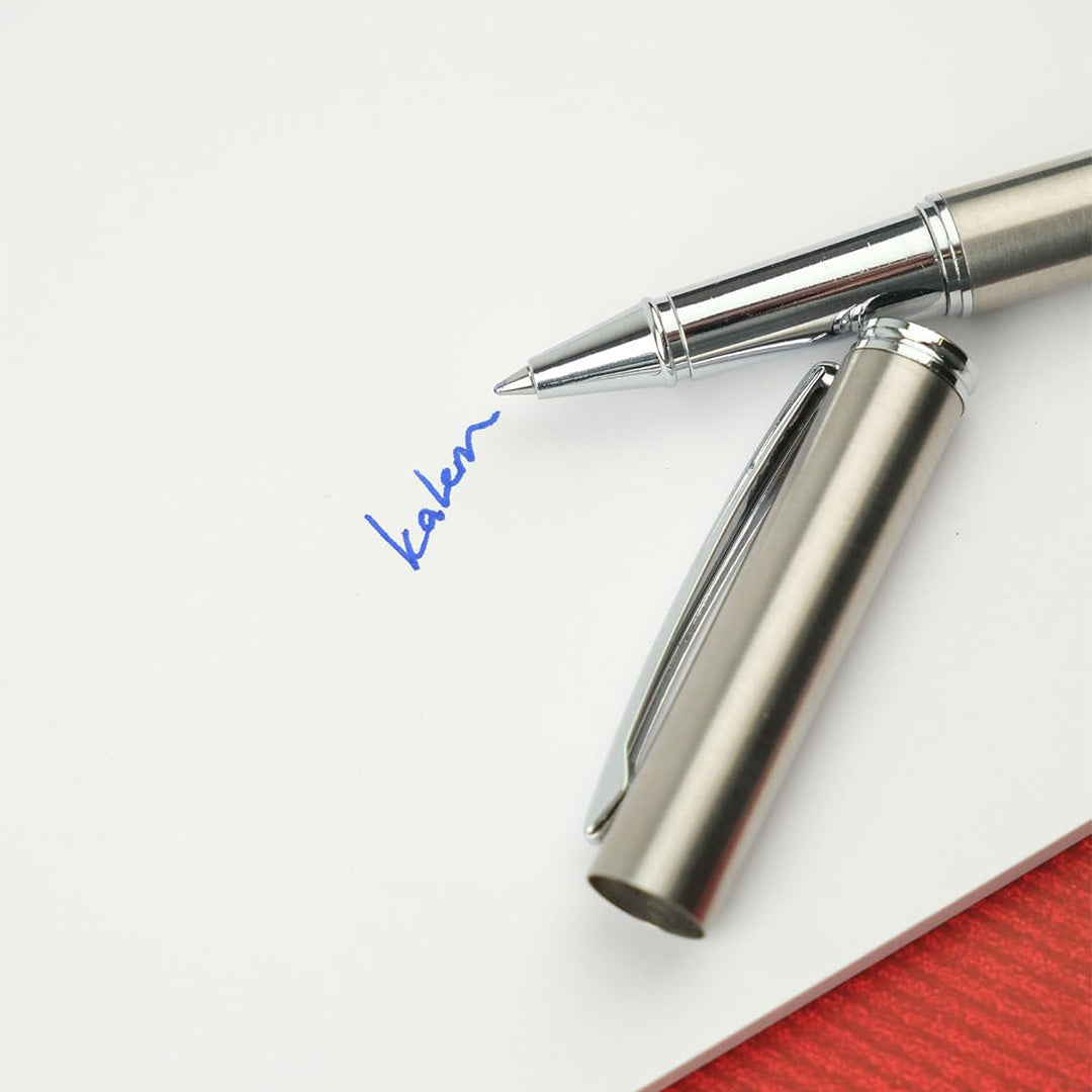 Scrikss 64 Series Rollerball Pen in Matte Chrome and its cap on a white surface with a red object partially visible.